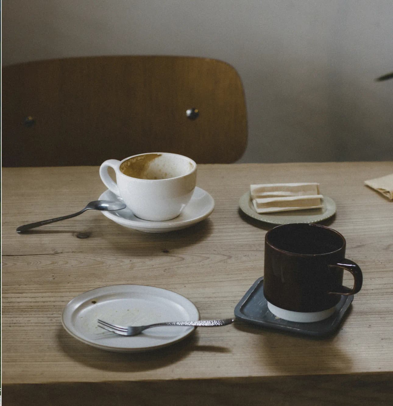 A finished cappuccino with a spoon on the saucer, a small plate with a fork, and a second mug on a weathered wooden table