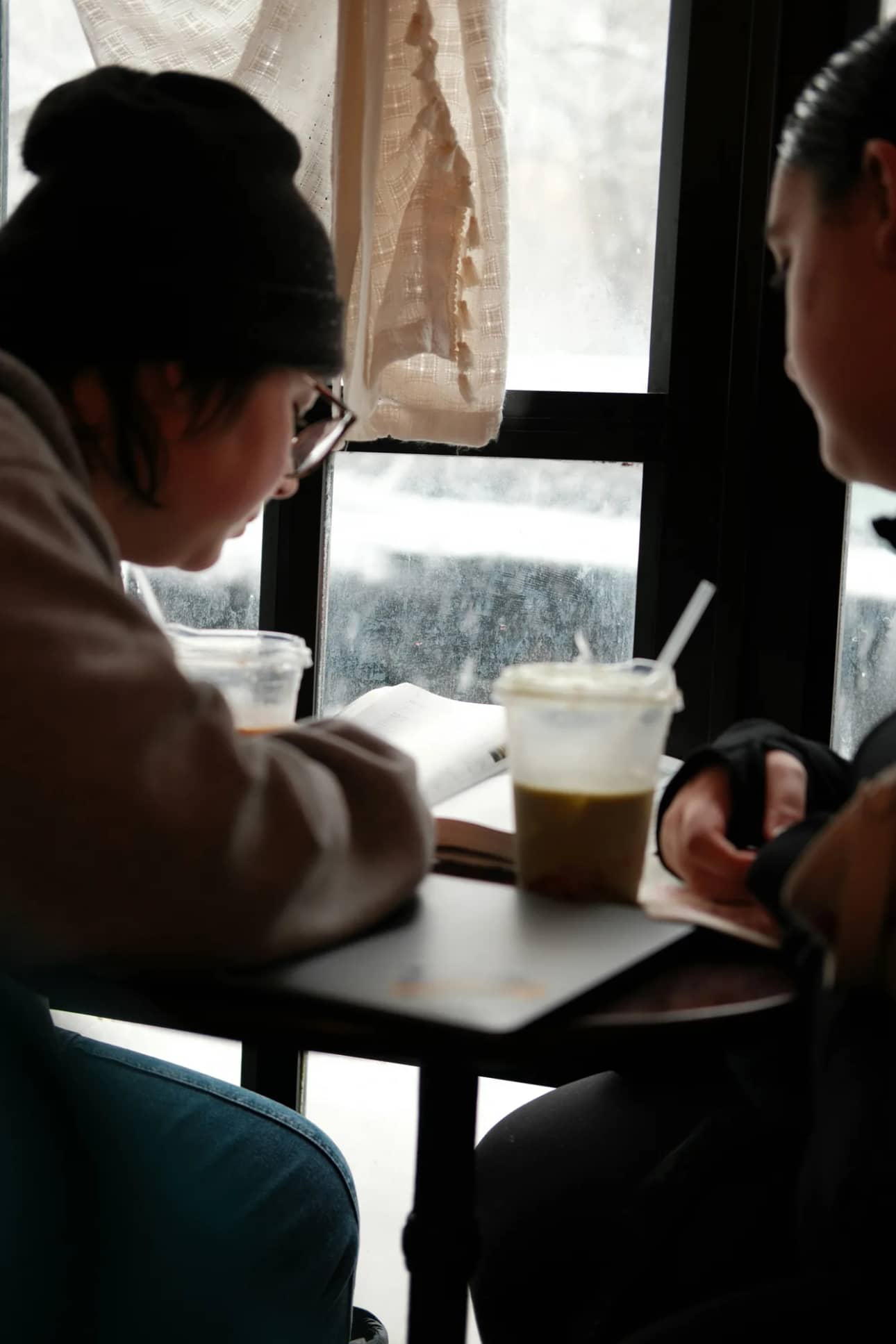 Two friends at a cafe window reading an open book together, warm winter light through frosted glass, iced drinks on the table