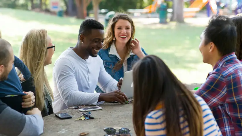 A group of friends gathered around a picnic table in a park, laughing mid-conversation, an open laptop between them