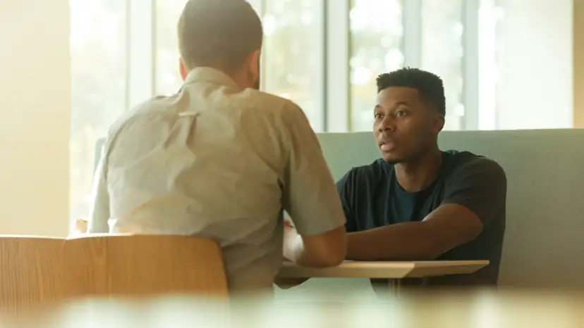 Two men across a booth table in earnest conversation, window light behind them