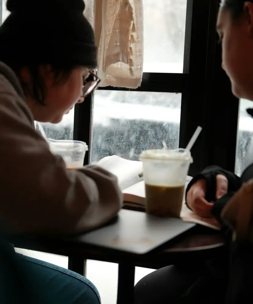 Two friends at a cafe window reading an open book together, warm winter light through frosted glass, iced drinks on the table